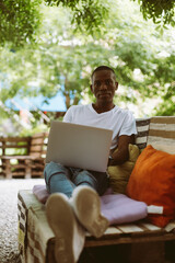 Obraz premium Vertical serious african american, dark skin man looking at camera, using laptop sitting on bench with legs in outdoor cafe. Prepare to online webinar, exams, e-learning education course with wireless