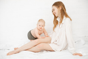 Portrait of young happy mother mom with long dark hair in white shirt with little cute plump baby sitting on white bed.