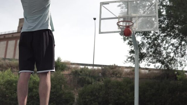 Dark-haired boy shooting a street basketball free throw. Image from behind a lonely young man scoring a two-point basket on a blue court. Sport concept
