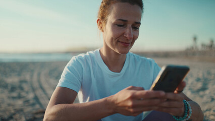 Close up woman checking her social media using smartphone by the sea. Sporty girl resting after workout on the beach