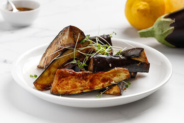 Grilled eggplant glazed with sauce, honey, microgreens on white plate and marble background, delicious vegan dish. Close up.