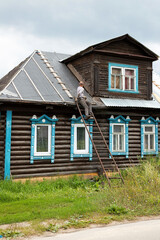 Fototapeta na wymiar Middle-aged man sitting back on metal ladder repairing roof of old wooden brown house carved windows