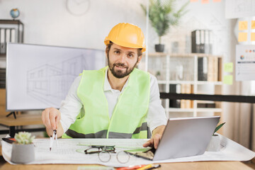 Young architect male in helmet and uniform sitting at table with blueprints and modern devices....