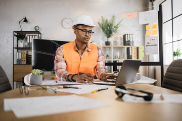 Young architect african male in helmet and uniform sitting at table with blueprints and modern devices. Focused man worker creating new building project sitting in modern office.