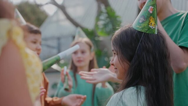 Birthday girl smiling while her friends congratulate her at a party on a sunny day. Green-themed birthday party. Boys and girls wearing birthday hats at the party. High quality 4k footage