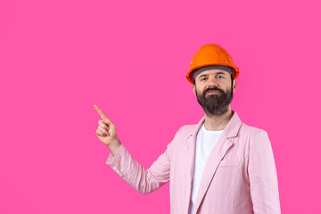 Portrait of young construction engineer wear orange hard hat, in a pink jacket standing on red studio background. The man points with his hand.