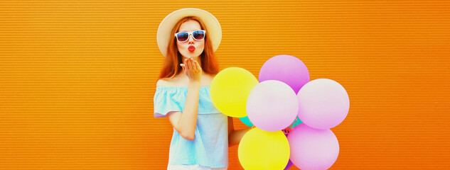 Summer portrait of happy woman blowing her lips with colorful balloons having fun wearing straw hat on orange background, blank copy space for advertising text