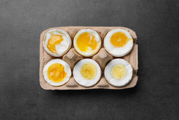 Boiled chicken eggs of different readiness stages in carton on dark grey table, top view