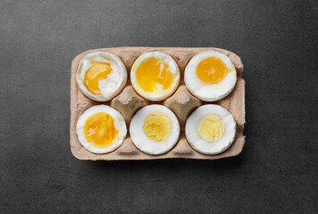 Boiled chicken eggs of different readiness stages in carton on dark grey table, top view