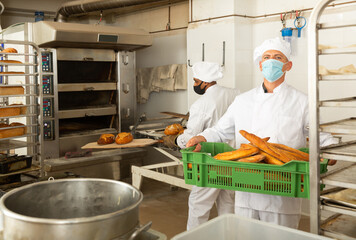 Baker in face mask for viral protection holding crate with bread, industrial kitchen of bakery on background