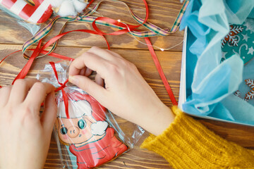 Girl's hands tie red ribbon on bag of gingerbread in form of pig in Santa Claus hat. Cookies with colored icing in box on wooden table. Christmas gifts. New Year's atmosphere.
