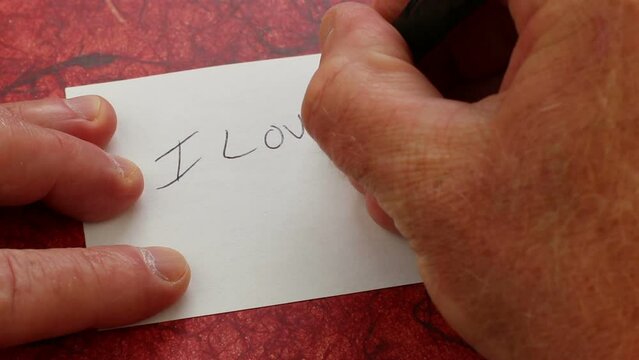 Thoughtful love note written printed by human hand fingers on a white index card in black ink close-up. White index card on a red abstract background fingers writing I love you with a smiley face