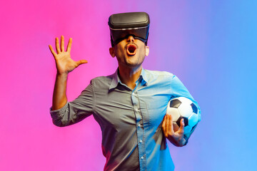 Portrait of excited man in shirt wearing virtual reality glasses and holding soccer ball, raised arm, supporting favorite football tram. Indoor studio shot isolated on colorful neon light background.