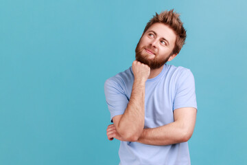 Portrait of smiling happy bearded man looking away with pensive expression, dreaming pleasant thoughts, fantasizing and making wish. Indoor studio shot isolated on blue background. - obrazy, fototapety, plakaty