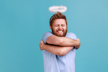 Portrait of bearded angelic man with holy nimbus standing smiling contentedly and hugging himself,...