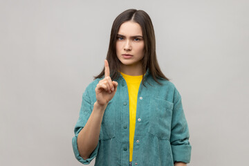 Be careful, Portrait of strict bossy young woman with brown hair seriously pointing finger up and warning, wearing casual style jacket. Indoor studio shot isolated on gray background.