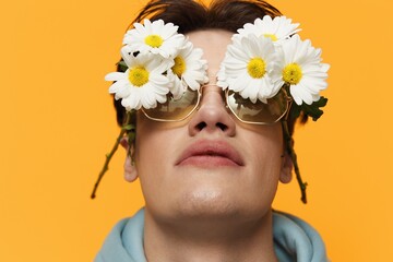 a close portrait of a handsome, funny man with daisies on his eyes, standing on a bright background in a light blue hoodie