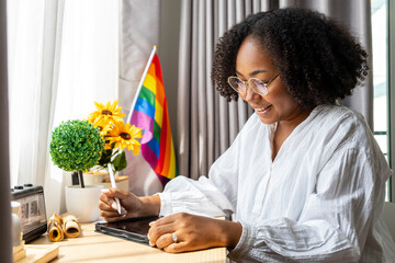 African American girl is working at home with LGBTQ rainbow flag in her table for coming out of closet and pride month celebration to promote sexual diversity and equality in homosexual orientation - obrazy, fototapety, plakaty