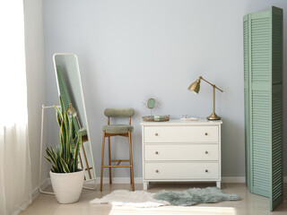 Stylish chest of drawers, chair, mirror and houseplant near light wall