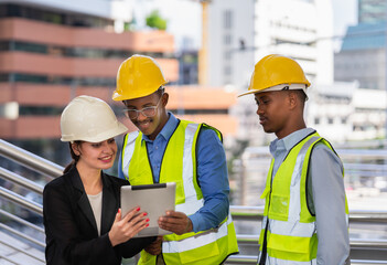 caucasian businesswoman in black suit wear helmet working on tablet with black engineer colleague. group of engineer working at construction site outside building in city