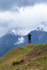 A tourist on a hill takes pictures of the beautiful snowy mountain peaks of the Eastern Sayan Mountains, covered with low clouds on a rainy day. Travel to the highlands, outdoor activities