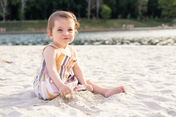 happy baby girl sitting on the beach on sand wearing striped summer dress on sunny day