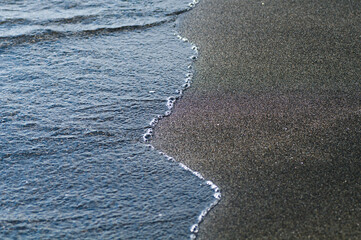 Black volcanic magnetic sand on the seashore. Background texture of the beach with waves closeup. 
