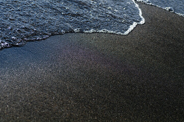 Black volcanic magnetic sand on the seashore. Background texture of the beach with waves closeup. 
