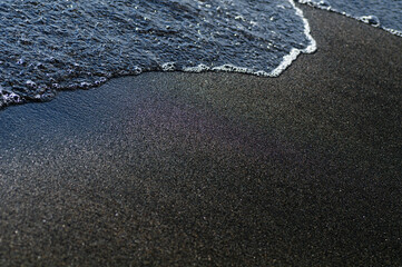 Obraz premium Black volcanic magnetic sand on the seashore. Background texture of the beach with waves closeup. 
