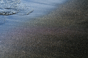 Black volcanic magnetic sand on the seashore. Background texture of the beach with waves closeup. 
