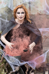 Fototapeta na wymiar Attractive beautiful woman in a Halloween costume black cloak and scary make-up. Young girl in a look of a witch dressed up for a party making fairy potion holding black lantern
