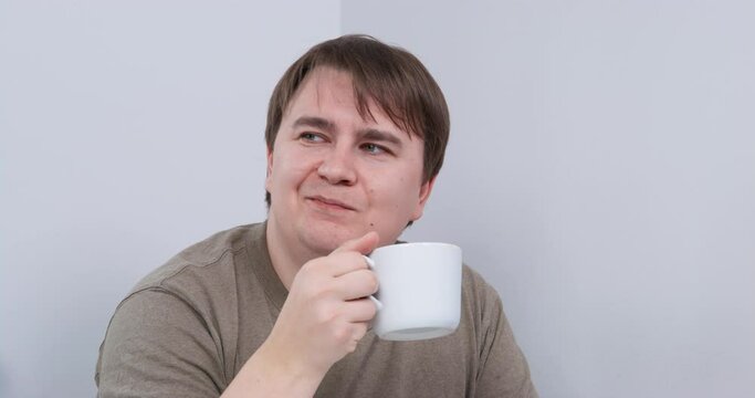 Portrait of young man with bracers, who is sitting at a table and drinking from a mug, front view. He enjoys the beverage, squints with pleasure and smiles after each sip