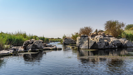 Fototapeta na wymiar A quiet backwater on the river. Picturesque boulders and green vegetation on the banks. Duckweed on the water. Reflection. Blue sky. Copy space. Egypt. Nile. Aswan