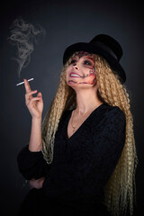 Beautiful Halloween Make-Up Style. Blond curly hair Model Wear Sugar Skull Makeup with Red Roses. Santa Muerte concept. Model on black background in studio with a cigar