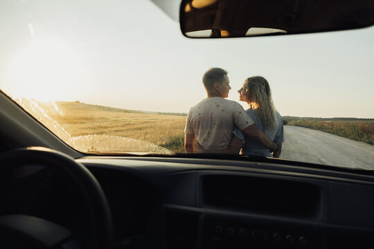 Back View Through Front Car Window on a Young Couple Hugging Near the Hood, Woman and Man Enjoying Road Trip at Sunset