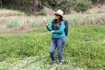 Latin adult woman with hat and sunglasses is in the middle of the forest using his cell phone to send and receive messages and search the location on the map	