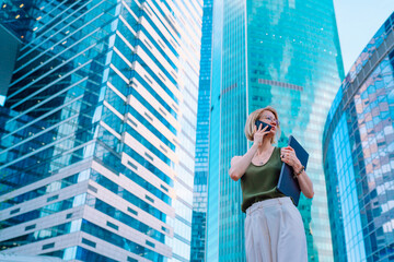 Portrait of busy middle-aged woman businesswoman with fair hair wearing green top, white trousers, standing on street in city centre near glass skyscrapers, holding blue laptop, talking on smartphone.