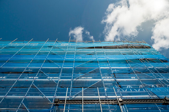 Building In Scaffolding And Blue Mesh Net Matching Blue Cloudy Sky. Construction And Renovation Industry In Town.