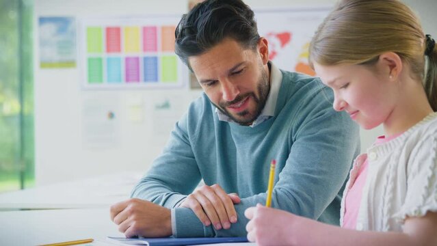 Male teacher sitting at desk with female elementary pupil in classroom as she writes in school exercise book - shot in slow motion
