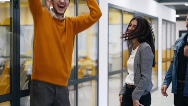 Young cheerful coworkers have fun during workday break. Two teams compete in skateboard racing in office hallway. Men pushing women on skateboards. Colleagues rejoice. Coworkers film on smartphones.