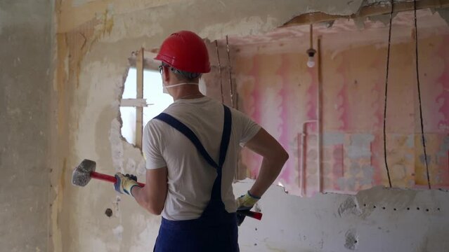 Contractor wrecks wall with sledgehammer making hole for rearrangement. Construction worker doing manual dismantling and demolition works with big hammer hits for apartment renovation in slow motion.