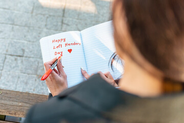 Young Woman lefty writes in a notebook. holds felt-tip pen in her left hand.Outdoors summer evening park. Left-handers Day August 13th.Closeup.
