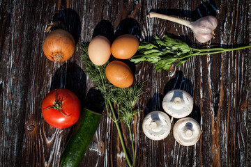 Creative composition of champignons, raw chicken brown eggs, cucumber, garlic, tomato, onion, decorated with herbs, dill and parsley, on a dark textured wooden background.