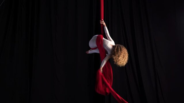 Professional circus performer. A flexible girl performs on stage on red aerial silks. Graceful curly-haired gymnast performs splits and gymnastic elements while dancing on canvases