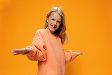beautiful. Darling. a cheerful girl of school age stands in an orange sweater on a yellow background raising her hands to the sides