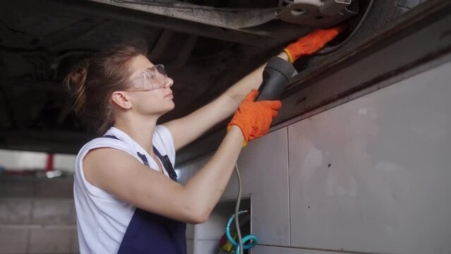 Female mechanic inspecting car suspension on hydraulic wheel play detector with remote control. Strong woman checks for wear in running gear and steering components. Service operator in overalls.