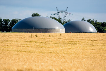 General view of a biogas plant with two digesters in a wheat field and an electricity pylon in the distance in the countryside.