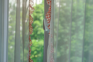Open pvc window frame with transparent silk curtain frilled with red ruche hanging at home room inside against blurred garden view
