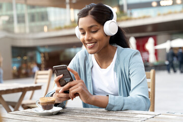 Smiling Indian woman holding  mobile phone shopping online, reading text message sitting in cafe. Happy asian hipster wearing white wireless headphones listening music using modern technology  