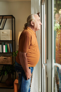 Pensive Senior Man With Long Hair Looking At Backyard Through Glass Entrance Door